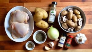 Ingredients for Chicken Mushroom Potatoes Crock Pot Recipe including chicken thighs, baby potatoes, mushrooms, onions, garlic, broth, cream, and herbs on a wooden countertop.