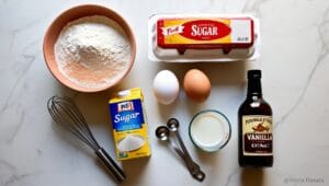 Flat lay of ingredients for funnel cake, including flour, eggs, sugar, milk, vanilla extract, whisk, and measuring spoons on a marble countertop