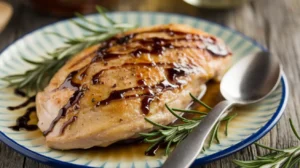 A golden-brown grilled chicken breast drizzled with honey balsamic glaze, garnished with fresh rosemary on a patterned plate