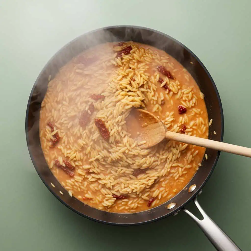 Orzo pasta cooking in broth with sun-dried tomatoes showing creamy sauce formation